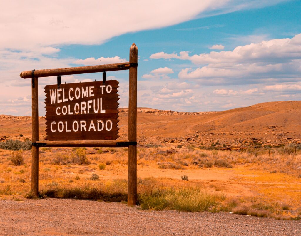 コロラド州カノンシティの入り口に立つ、木製の「WELCOME TO COLORFUL COLORADO」と書かれた看板。背景には広大な乾燥した砂漠地帯と青空が広がっている。